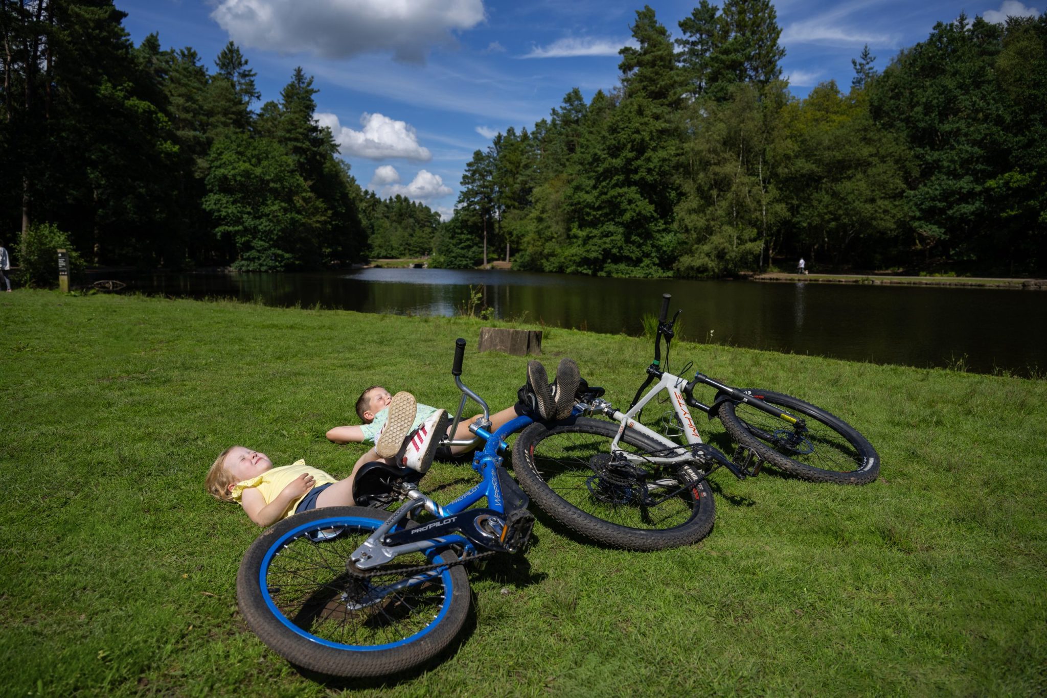 Little girl and boy lying down sunbathing next to mallards pike lake resting their feet up on bike and tagalong that is lying on the ground